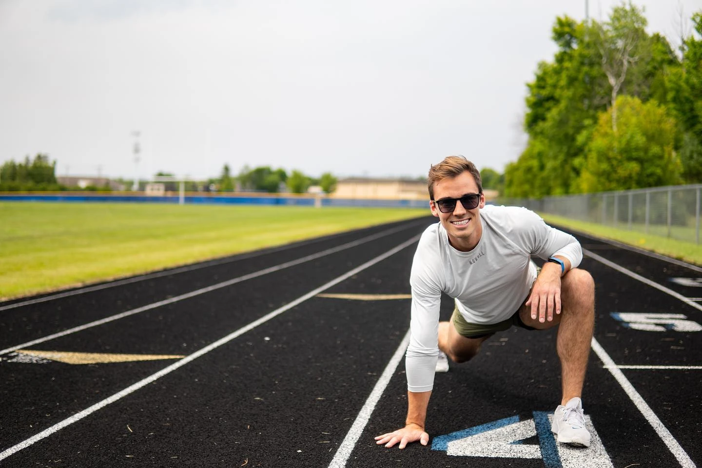 Sam Leicht in sunglasses and a long-sleeve top kneeling on one knee on a running track, smiling at the camera