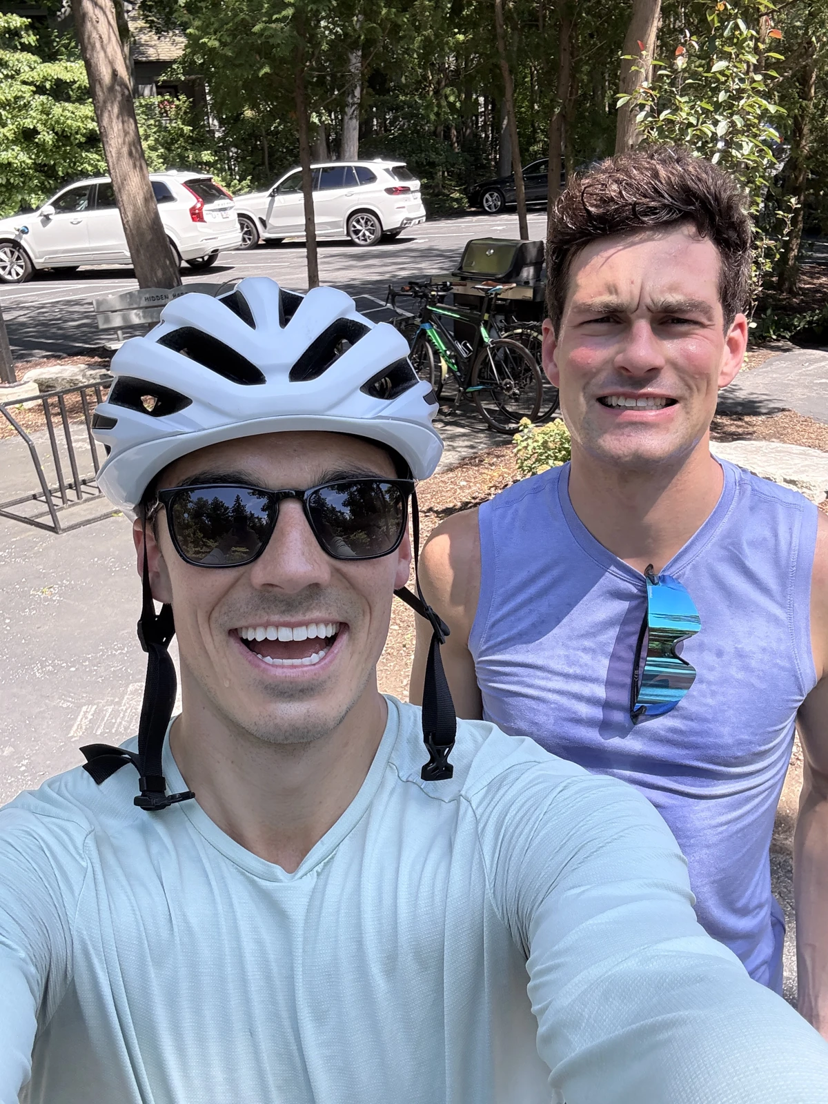 Sam Leicht in a white bike helmet taking a smiling pre-race selfie with another person outdoors, with bikes parked behind them