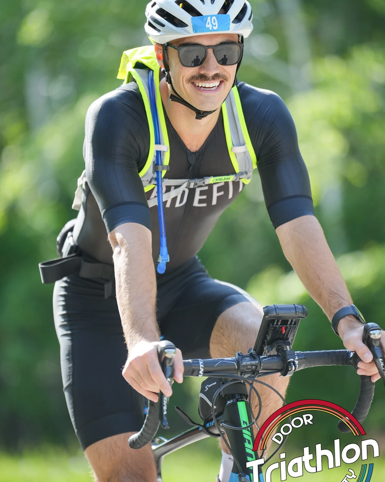 Sam Leicht smiling on a road bike in a white helmet with race number 49 and a Pridefit tri suit, with a Door County Triathlon watermark
