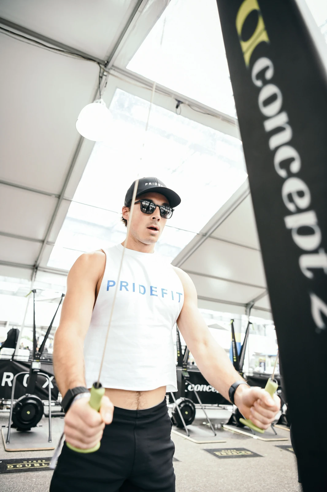Sam Leicht in a Pridefit cap, sunglasses and tank pulling the handles of a Concept2 ski erg inside a HYROX race tent