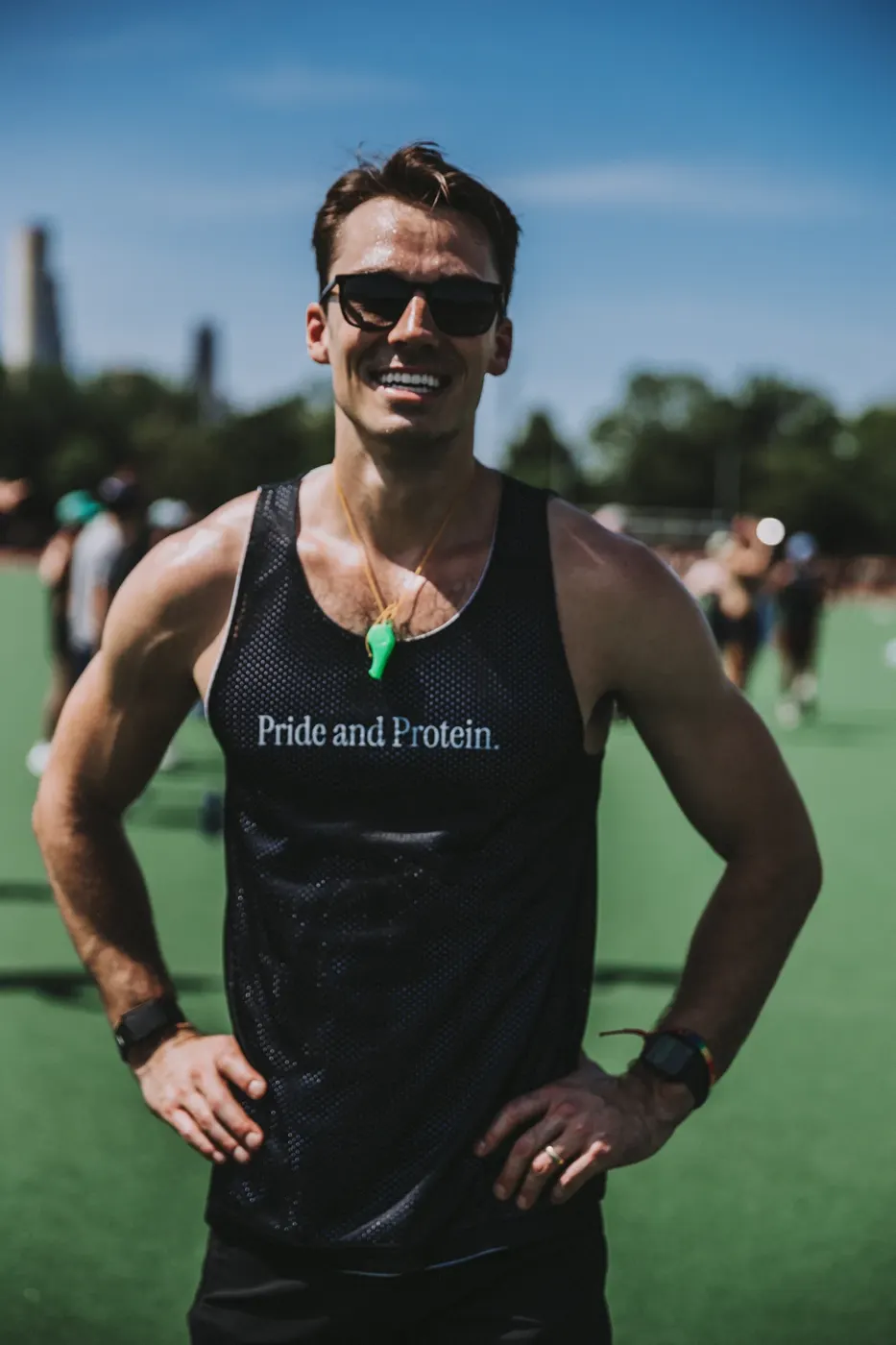 Sam Leicht grinning in sunglasses and a 'Pride and Protein' tank at an outdoor Pride workout, with participants on the field behind him