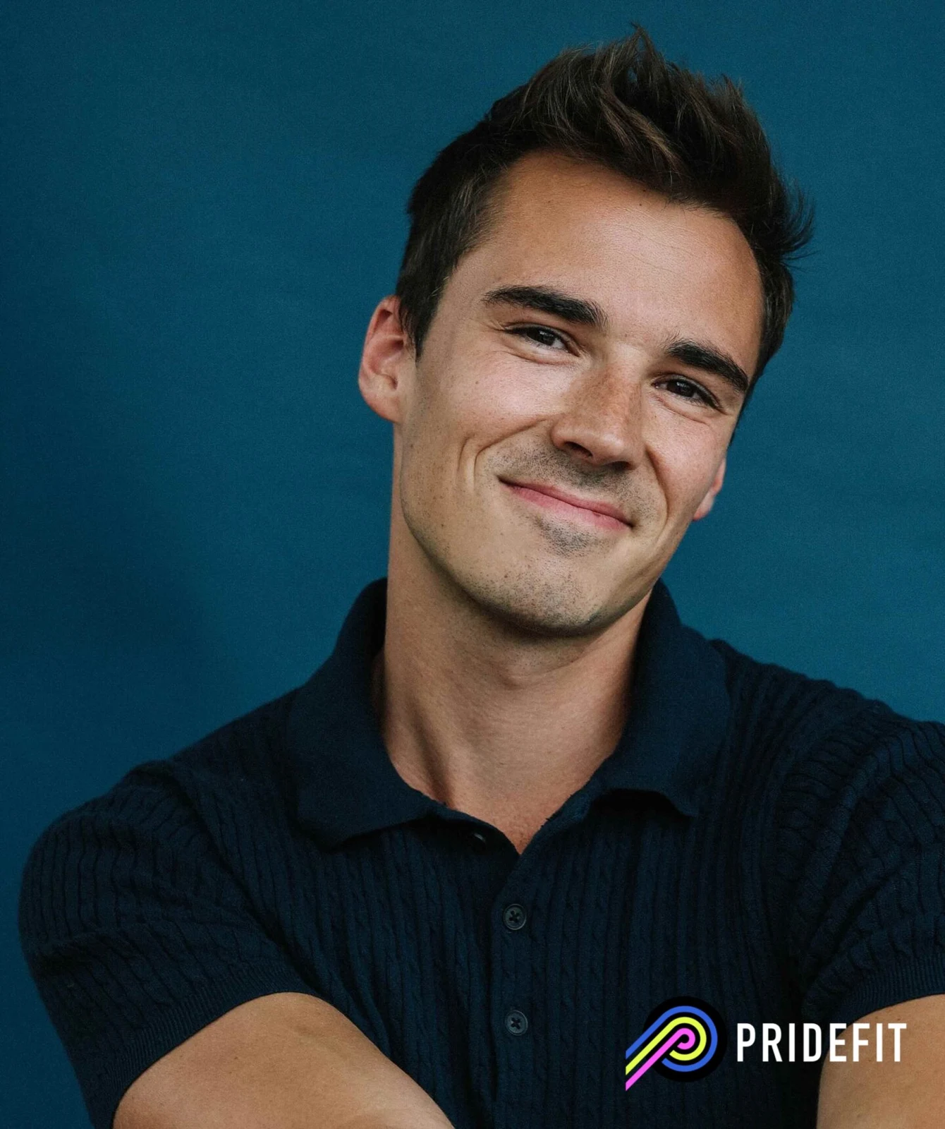 Sam Leicht smiling in a navy knit polo against a blue backdrop, with the Pridefit logo in the corner