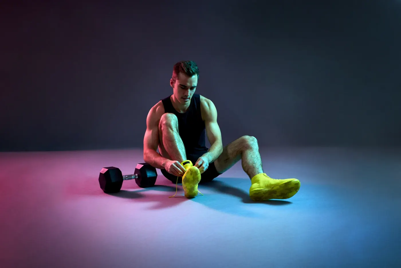 Sam Leicht sitting on a studio floor tying neon yellow training shoes beside a dumbbell, lit in pink and blue