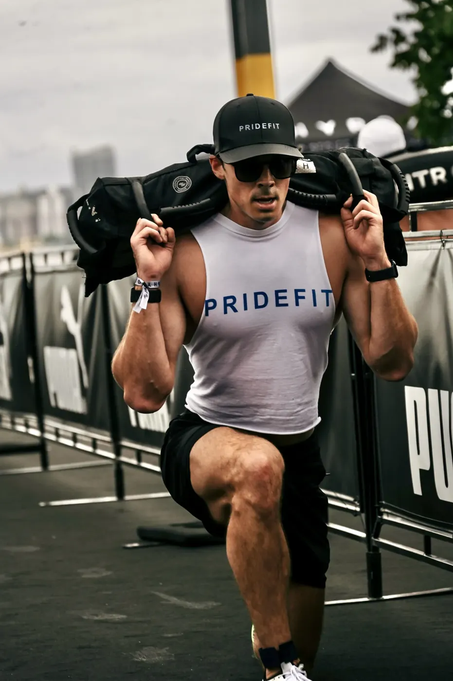 Sam Leicht in a Pridefit cap and tank doing a walking lunge with a sandbag across his shoulders at a fitness competition