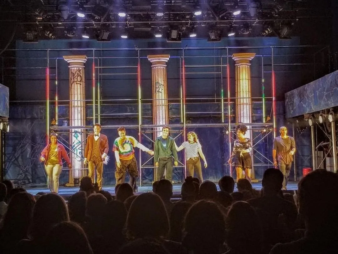 A musical company taking a curtain call beneath tall columns on a theater stage