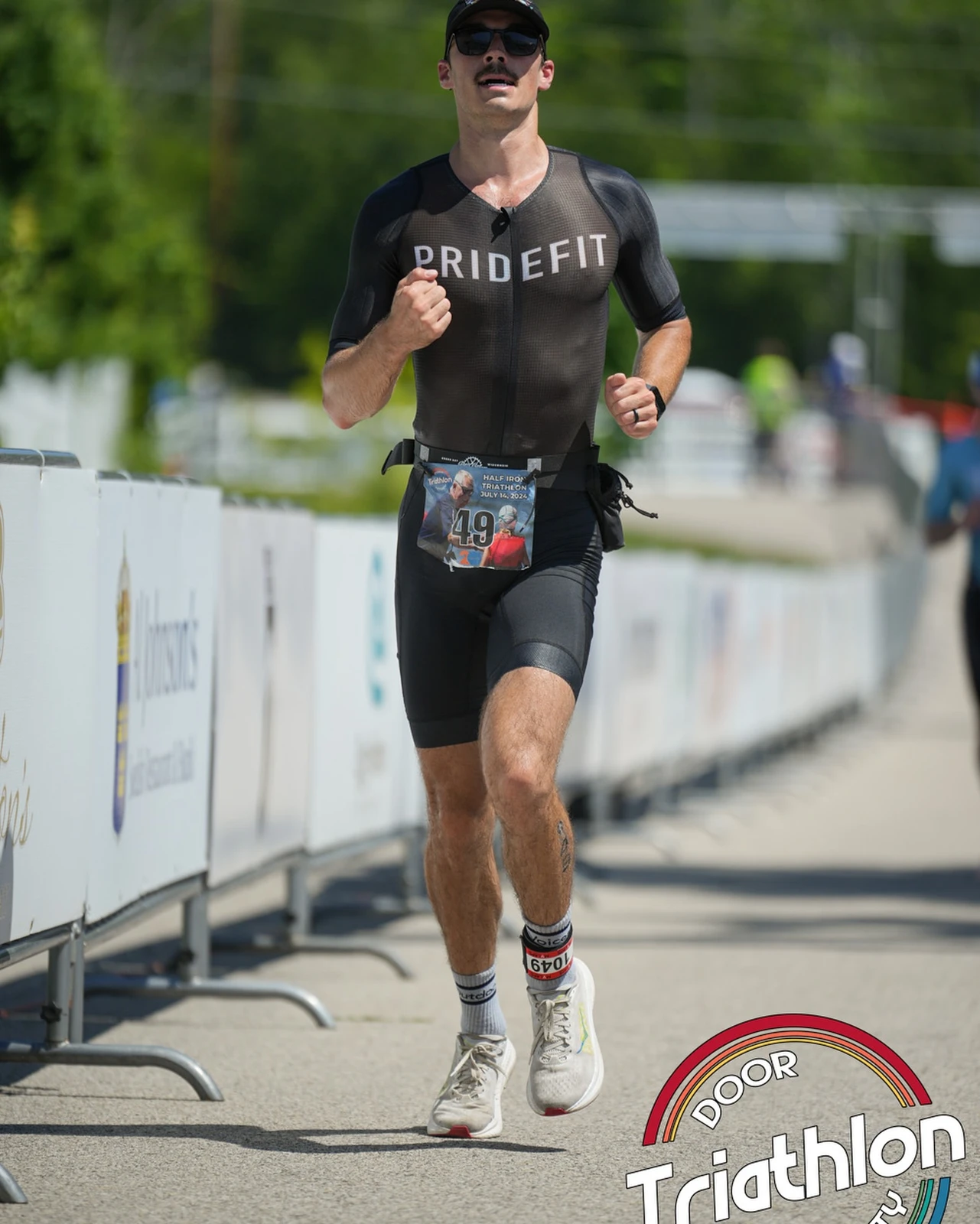 Sam Leicht running beside the barriers in a black Pridefit tri suit wearing race bib 49, with a Door County Triathlon watermark
