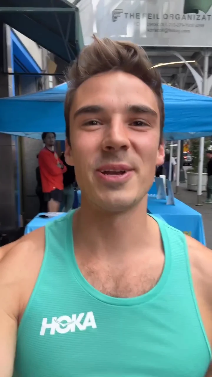 Sam Leicht in a teal HOKA tank talking to camera on a New York sidewalk beside a blue event tent